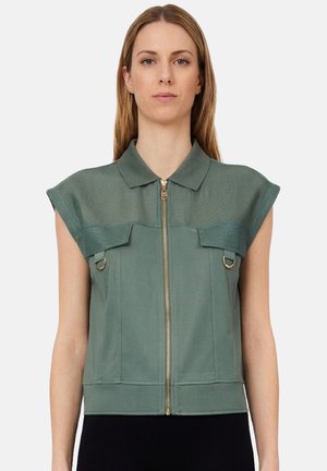 Donna con capelli biondi lisci che indossa un top verde senza maniche con cerniera, colletto largo e anelli decorativi in metallo sul petto.