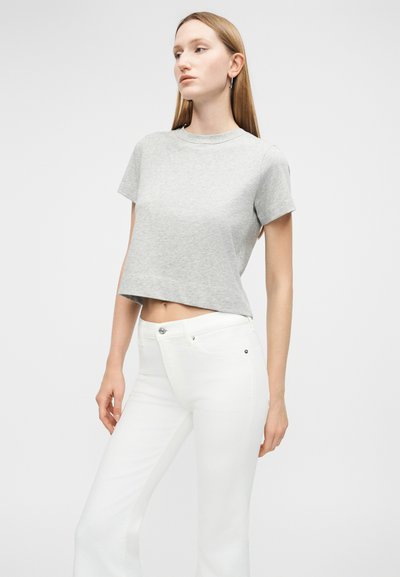 Γυναίκα με ίσια ξανθά μαλλιά που φοράει ανοιχτό γκρι κοντομάνικη μπλούζα και ψηλόμεσα λευκά παντελόνια, στέκεται με το ένα χέρι λυγισμένο.