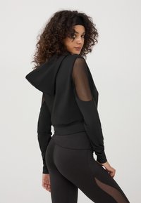 Felpa sportiva nera con un ampio cappuccio, maniche in rete trasparente e design aderente; abbinata a leggings neri a vita alta con ritagli in rete.