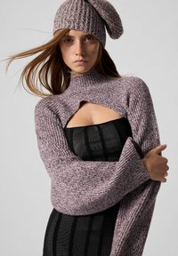Kvinde iført en lavendelstrikket hat og en matchende cropped sweater over en stropløs sort kjole, stående foran en ensfarvet lys baggrund.