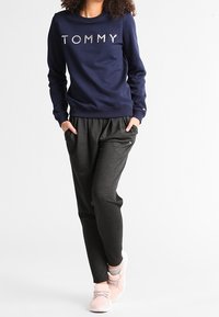 Felpa blu navy con "TOMMY" in lettere argentate, abbinata a pantaloni jogger grigi scuri con tasche laterali e sneakers rosa chiaro.
