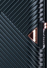 Surface texturée noire d'une valise avec des rainures diagonales et une plaque logo triangulaire en métal rose doré près d'une serrure TSA.