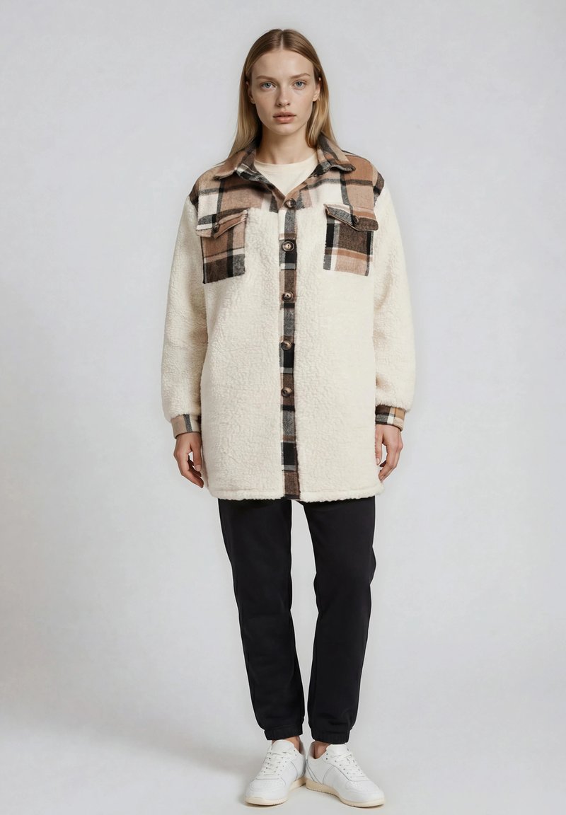 Femme portant une veste sherpa crème avec des accents à carreaux marron, un pantalon noir et des baskets blanches, debout devant un fond gris uni.