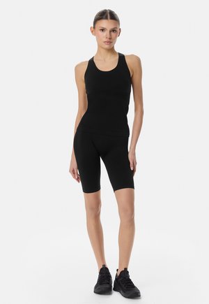 Naine seisab seljas musta värvi varrukateta kitsas topp, mustad põlveni ulatuvad retuusid ja mustad spordijalatsid, taust on helevalge.