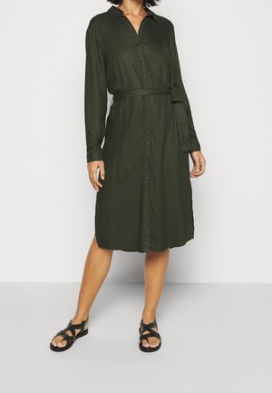 Kobieta ubrana w oliwkową, zapinaną na guziki sukienkę midi z długim rękawem, z dopasowanym paskiem w talii i czarnymi sandałami z ćwiekami.