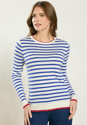 Mujer con suéter blanco de manga larga con rayas horizontales azules y ribete rojo, combinada con jeans azul oscuro, de pie frente a un fondo liso.