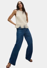 Top senza maniche bianco con texture e orlo frangiato, abbinato a jeans a gamba larga blu scuro e ciabatte beige. Un look semplice e informale.