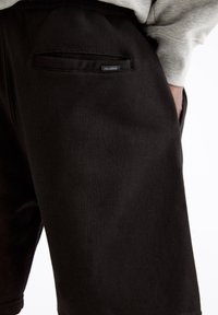 Primo piano di pantaloni corti neri con tasca posteriore e piccolo etichetta Pull&Bear, indossati con un sudore grigio, mano poggiata nella tasca laterale.