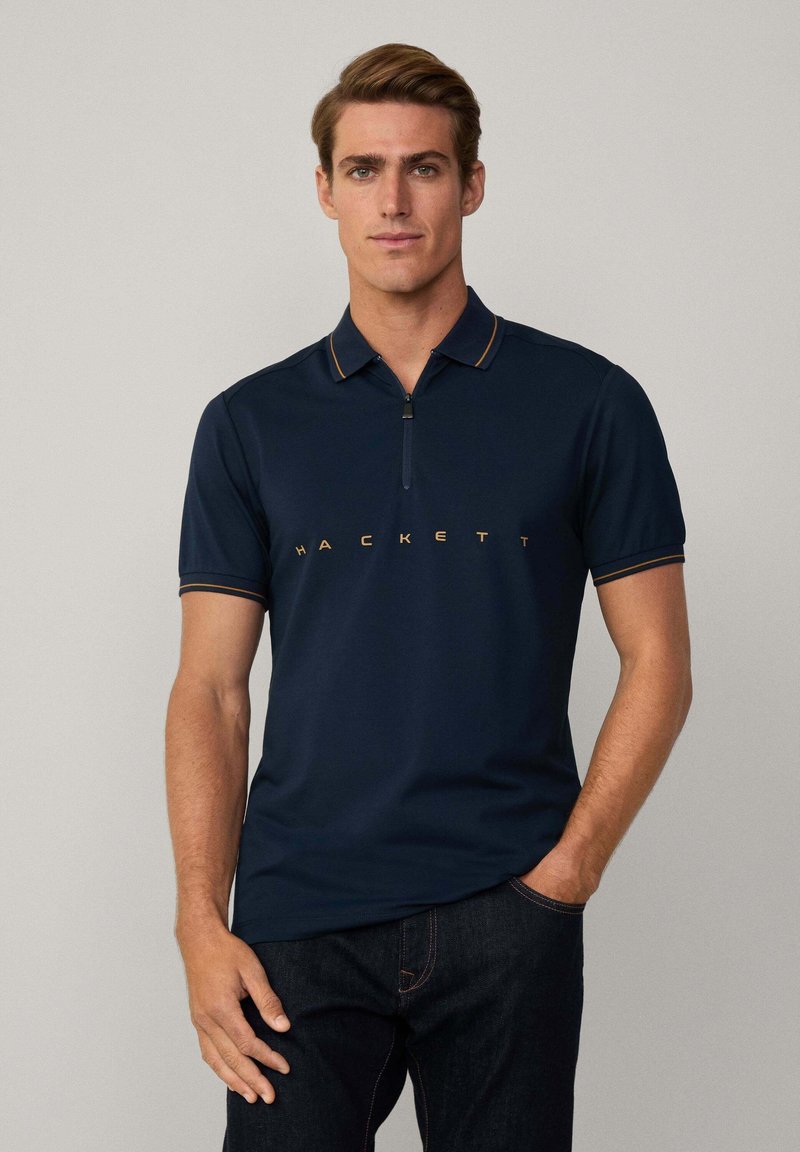 Camisa polo de color navy con mangas cortas, cuello con cremallera y ribete naranja, que presenta el logo "HACKETT" en letras grandes en el pecho.