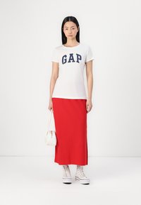 Balta medvilninė marškinė su tamsiai mėlynu "GAP" logotipu, derinama su ilgu raudonu sijonu. Balti sportbačiai ir balta rankinė užbaigia įvaizdį.