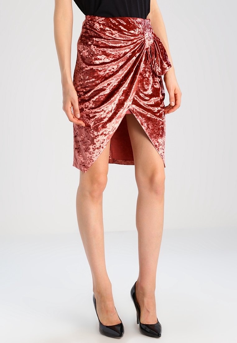 Jupe portefeuille en velours rose poudré avec un nœud sur le côté, une fente haute et une texture douce. Associée à des talons hauts noirs.