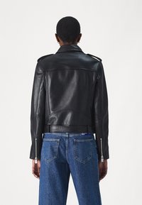 Crna kožna moto jakna s uskim dizajnom, epauletama na ramenima i zip-zatvaračima na rukavima, u kombinaciji s svijetloplavim trapericama.