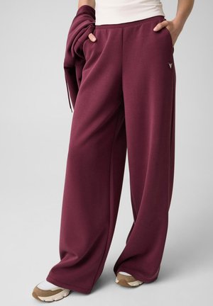 Personne portant un pantalon large bordeaux et des baskets beige, debout les mains dans les poches devant un fond uni.