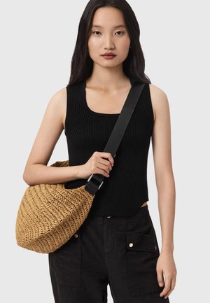 Femme portant un haut sans manches noir et un pantalon, tenant un sac à bandoulière tissé beige avec une large sangle noire sur la poitrine.