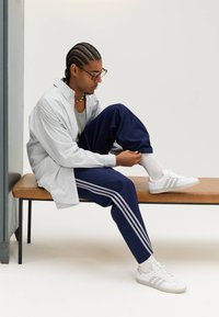 Prugasta košulja na kopčanje, sivi top bez rukava, mornarski hlače s bijelim prugama, bijele tenisice i ogrlica od perlica. Osoba sjedi na klupi.