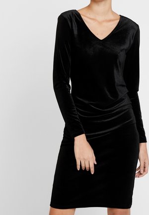 Frau trägt ein schwarzes Samtkleid mit langen Ärmeln und V-Ausschnitt, tailliert, steht vor einem schlichten hellgrauen Hintergrund.