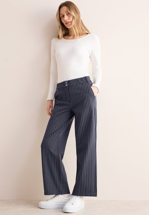 Pantalones de pierna ancha a rayas en tejido azul marino con rayas blancas, con una cinturilla abotonada, bolsillos laterales y combinados con zapatos blancos.
