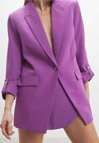 Lila skräddarsydd kavaj med djup v-ringning och uppvikta ärmar. Har två framfickor och en enkel knappstängning. Slen textur.