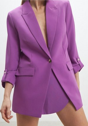 Violet bleiseri, joka on räätälöity syvästä V-aukosta ja käärityistä hihoista. Siinä on kaksi etutaskua ja yksi nappikiinnitys. Sileä rakenne.