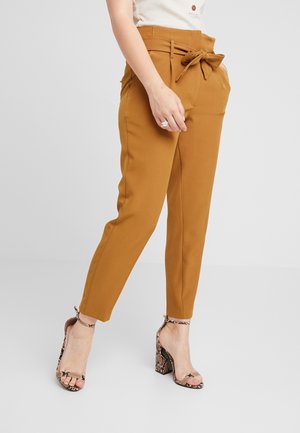 Vrouw draagt mosterdkleurige paperbag-broek met een gestrikte riem en slangenprint hoge hakken sandalen, hand rust langs het lichaam.