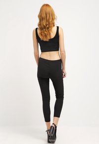 Kvinna med långt vågigt rött hår, iförd en svart ärmlös kort topp, svarta tajta byxor och svarta högklackade skor, står vänd bort.