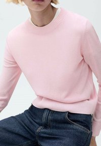 Jasnoróżowy sweter z dzianiny z okrągłym dekoltem i ściągaczami, zestawiony z ciemnoniebieskimi jeansami. Miękka struktura, dopasowany krój.