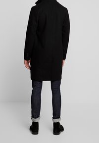 Schwarzer Wollmantel mit taillierter Passform, hohem Kragen und sichtbaren Nähdetails. Kombiniert mit dunklen Jeans und schwarzen Wildleder-Stiefeletten.