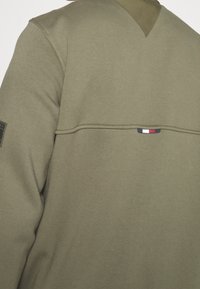 Bluza w kolorze oliwkowej zieleni wykonana z miękkiej tkaniny, o fakturze dzianiny, z opuszczonymi ramionami i małym logo w kolorach czerwonym, białym i niebieskim.