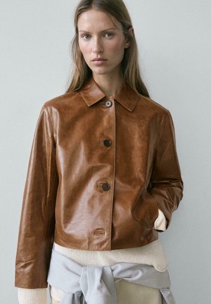 Femme portant une veste boutonnée en cuir marron par-dessus un pull en maille, avec une veste gris clair nouée autour de la taille, posant devant un fond uni.