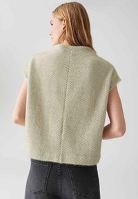 Femme aux cheveux blonds portant un pull sans manches de couleur crème texturé et un jean gris foncé, de dos contre un fond uni.