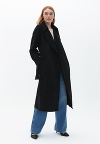 Cappotto lungo nero in tessuto morbido con colletto a punta, apertura frontale e tasche laterali, abbinato a jeans larghi azzurri e scarpe nere.