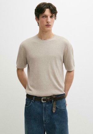 Junger Mann trägt einen beigen kurzärmligen Strickpullover, blaue Jeans mit einem dunklen geflochtenen Gürtel, steht vor einem schlichten weißen Hintergrund.