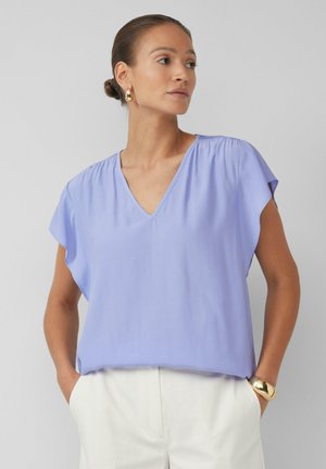 Kurzärmlige lavendelfarbene Bluse mit V-Ausschnitt, weichem Stoff und dezentem Raffung an den Schultern. Kombiniert mit weißen Hosen.