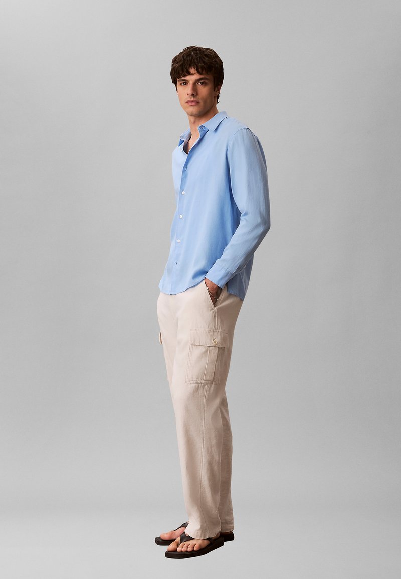Jonge man staand zijwaarts, draagt een lichtblauw overhemd met lange mouwen, beige cargobroek en zwarte slippers tegen een effen grijze achtergrond.