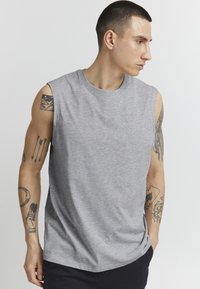 Graues ärmelloses T-Shirt aus weichem Material, mit einem runden Halsausschnitt und einem lässigen Schnitt, wobei die sichtbaren Tattoos an den Armen des Modells zu sehen sind.
