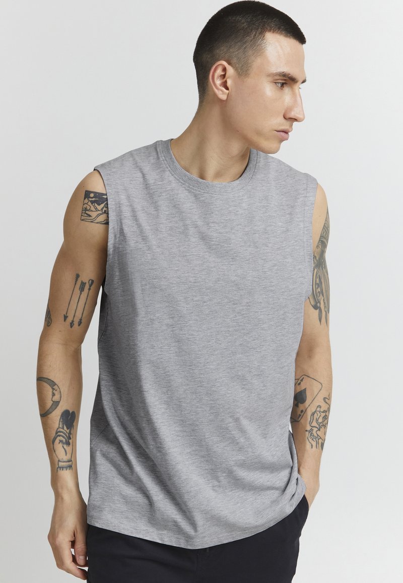 Graues ärmelloses T-Shirt aus weichem Material, mit einem runden Halsausschnitt und einem lässigen Schnitt, wobei die sichtbaren Tattoos an den Armen des Modells zu sehen sind.