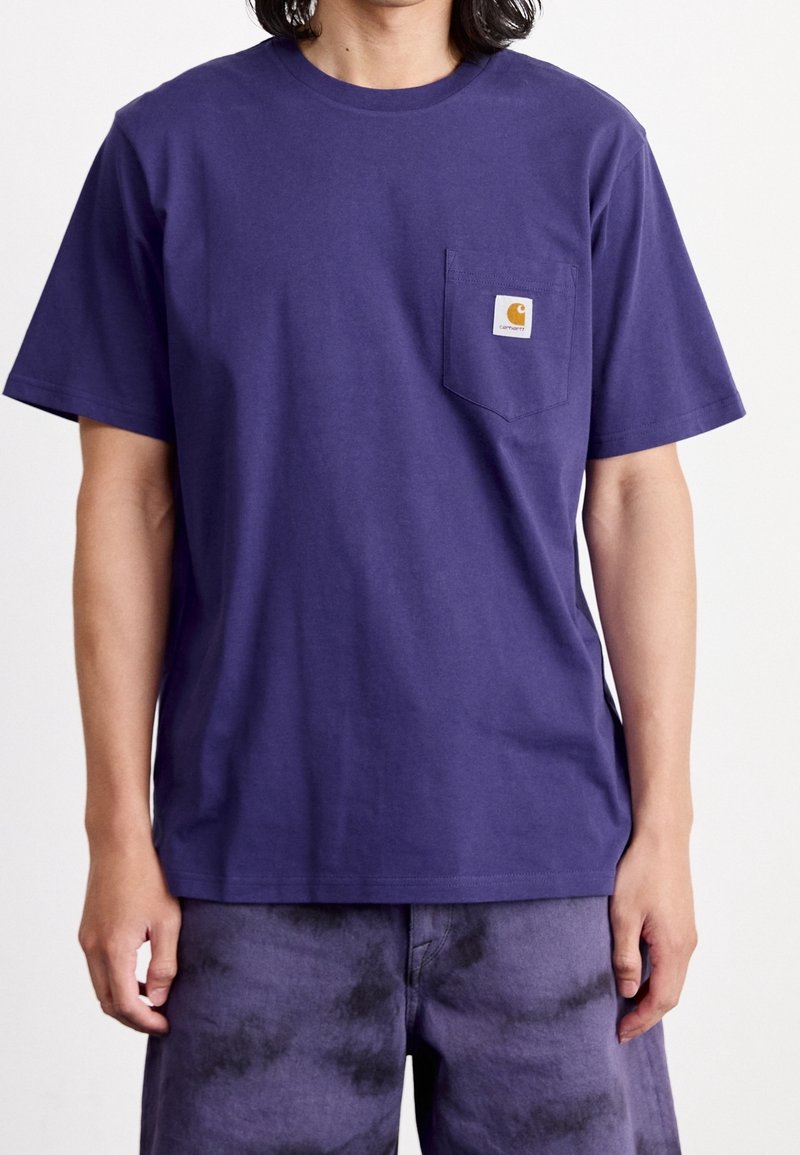 T-shirt di cotone viola con scollo rotondo e maniche corte, dotato di taschino sul petto con etichetta del logo. Il tessuto appare liscio e senza trama.
