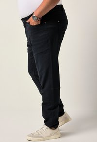 Uomo che indossa pantaloni neri, sneakers bianche, una camicia bianca infilata dentro i pantaloni e un orologio da polso con un cinturino a righe nere e rosse, in piedi di lato.
