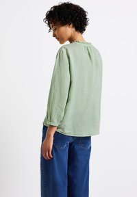 Camicia verde chiaro a maniche lunghe con scollo tondo e dettaglio arricciato sul retro, abbinata a jeans blu larghi. Tessuto in lino morbido.