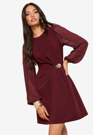 Bordeauxrode jurk met een getailleerd lijfje en een uitlopende rok, voorzien van doorschijnende pofmouwen en een decoratieve gouden knoop in de taille.