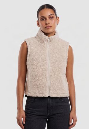 Femme portant un gilet polaire beige sans manches avec une fermeture éclair frontale et un pantalon noir, debout devant un fond clair uni.