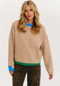 Jasnobrązowy, oversize sweter z niebieskimi i zielonymi prążkowanymi akcentami przy dekolcie i dolnym obszyciu, w połączeniu z spodniami w panterkę. Miękka tekstura.