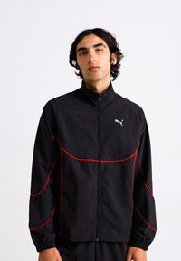 Svart treningsjakke med røde detaljer; har høyt krage, glidelås foran og Puma-logo. Laget av lett materiale med en jevn tekstur.