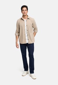 Camisa beige de botones con rayas verticales, camiseta blanca por debajo, vaqueros oscuros y zapatillas blancas. El modelo está de pie contra un fondo blanco.