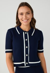 Navy gestrickte kurzärmelige Strickjacke mit einem weißen Kragenhals, durchgehenden Knöpfen und horizontalen weißen Streifen an den Ärmeln und am Saum.