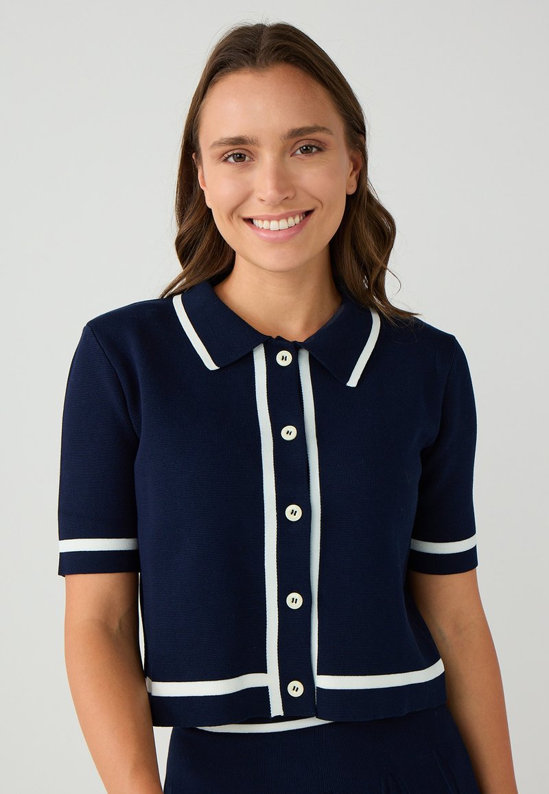 Navy gestrickte kurzärmelige Strickjacke mit einem weißen Kragenhals, durchgehenden Knöpfen und horizontalen weißen Streifen an den Ärmeln und am Saum.