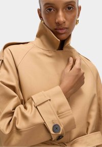 Beiger Trenchcoat mit hohem Kragen, langen Ärmeln und Knopfdetails an den Bündchen. Glatte Textur und modernes Design.