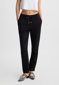 Pantaloni jogger neri a gamba tapered con vita elastica, coulisse e accessori in tonalità oro. Indossati con un top bianco corto e scarpe con motivi.