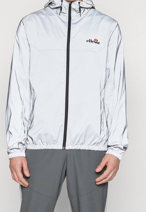 Mann trägt eine glänzende silberne Kapuzenjacke mit Reißverschluss, elastischen Bündchen und Saum sowie graue Hose. Die Jacke hat ein kleines ellesse-Logo auf der Brust.