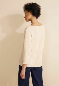 Maglione rosa chiaro con una maglia strutturata, scollo tondo e maniche a 3/4, abbinato a jeans blu scuro. Design minimalista senza motivi visibili.